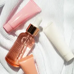 Un frasco de serum con gotero y tubos de crema rosa, blanco y melocotón sobre tela blanca. ¿Que va primero el serum o la crema?