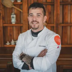 Carlos Maldonado, chef con tatuajes y barba, sonríe con los brazos cruzados frente a una estantería de madera.
