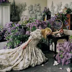 Una mujer rubia con un vestido estampado yace rodeada de lilas, una escena onírica capturada por Tim Walker.