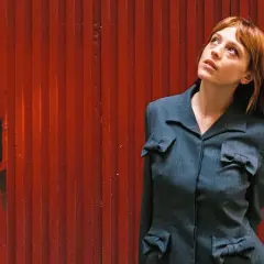Catalina Sopelana, con su cabello rojizo y mirada perdida, posa frente a una puerta roja acanalada, luciendo un traje gris con detalles de lazos.
