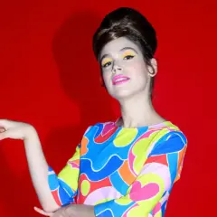 Cosima Ruiz de la Prada, con un peinado voluminoso y maquillaje vibrante, posa con un vestido de estampado psicodélico sobre fondo rojo.