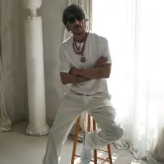 Pierpaolo Piccioli, con gafas de sol y collares llamativos, posa con los brazos cruzados en un entorno elegante con cortinas y columnas.