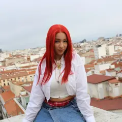 Joven con cabello rojo vibrante, como una rosa encendida, posa sobre tejados urbanos.