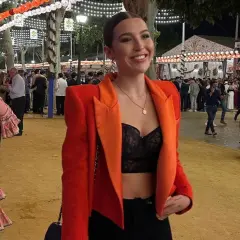 Chica sonriente con chaqueta naranja y top negro, lista para la ropa para ir a la feria de dia. Ambiente festivo con luces y gente.