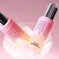 Dos frascos de aceite para pelo, con un degradado rosa y amarillo, reposan sobre cubitos de hielo en una copa.