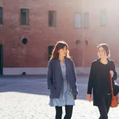 Las hermanas Arce caminan juntas bajo el sol, conversando animadamente frente a un edificio de ladrillo.