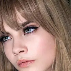 Primer plano de una mujer con flequillo rubio desfilado, delineado de ojos dramático y labios rosados.