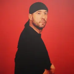 Retrato de Manuel Piña con gorro y camiseta negra, sobre fondo rojo vibrante.
