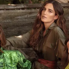 Belen Corsini y dos mujeres más, vestidas con ropa de tonos verdes, posan sentadas en un banco de madera rústico.