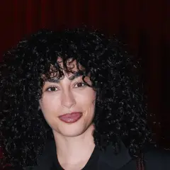 Mina El Hammani, con su cabello rizado y oscuro, sonríe ante un fondo de cortinas rojas.