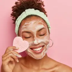 Mujer sonríe con espuma en la cara, usando una esponja en forma de corazón para su limpieza facial.