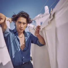 Enzo Vogrincic, con su camisa desabrochada y collar llamativo, posa entre sábanas blancas tendidas al sol.