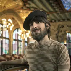 David Ucles sonríe con un gorro y una camiseta a rayas, en un salón con vidrieras y lámparas doradas.