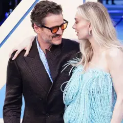 Pedro Pascal y su pareja, con ella luciendo un vestido azul con flecos, se miran tiernamente en una alfombra azul.