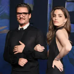Pedro Pascal y una mujer elegante posan juntos. El actor, con gafas y un suéter negro, luce un cabestrillo. Ella, con un vestido negro y joyas brillantes, sonríe.