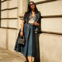 Mujer con gafas de sol posa apoyada en una pared, luciendo un conjunto vaquero y una falda vaquera larga.