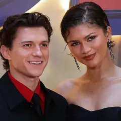Tom Holland y Zendaya sonríen juntos en un evento. Él lleva una camisa roja y corbata negra. Ella luce un vestido negro y pendientes largos.