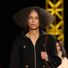 Una mujer con cabello rizado y gris camina por una pasarela, luciendo un elegante cárdigan negro con botones dorados, como una **chanel cantante**.
