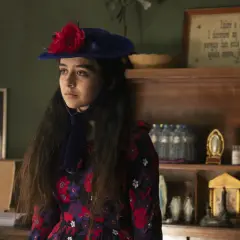 Irene Balmes, con un sombrero azul y un vestido floral, mira pensativa. Detrás, estanterías con objetos religiosos y un cuadro.