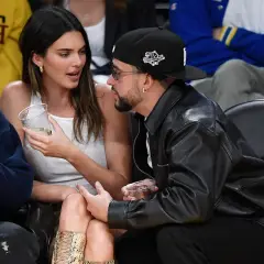 Bad Bunny y Kendall Jenner conversan en un evento deportivo, ella con un vaso en la mano y él con chaqueta de cuero.