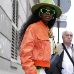 Mujer con sombrero verde y gafas neón, luciendo una bomber naranja vibrante.