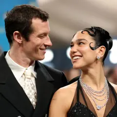 Dua Lipa y su novio, Romain Gavras, sonríen mirándose. Ella luce un vestido negro y joyas deslumbrantes.