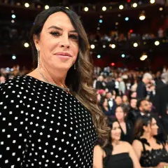 Karla Sofía Gascón, con un vestido negro de lunares blancos, sonríe en un evento con público al fondo.
