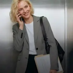 Mujer sonriente en ascensor, con un elegante outfit oficina, hablando por teléfono y cargando carpetas.