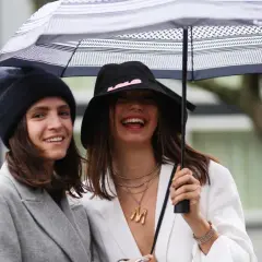 Dos mujeres ríen bajo un paraguas, luciendo un estiloso outfit lluvia mujer con gorros y abrigos.