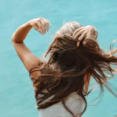 Mujer con cabello largo y castaño, con algunas canas visibles, moviendo su pelo al viento. Una alternativa natural para cubrir canas sin tinte.