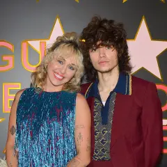Miley Cyrus y Max Morando posan sonrientes. Ella lleva un vestido azul brillante con flecos, él un traje burdeos con detalles dorados.