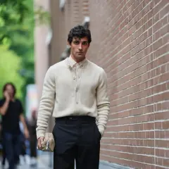 Hombre con suéter crema y pantalón negro, bolso de mano beige. Un ejemplo de las últimas tendencias moda hombre.