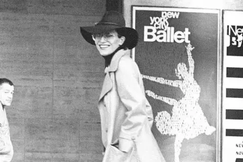 Elsa Peretti, con sombrero y gafas, sonríe frente a un cartel del New York City Ballet.