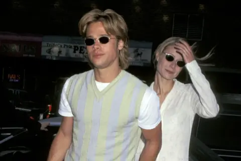 Brad Pitt joven con gafas de sol y un suéter a rayas, junto a una mujer rubia.