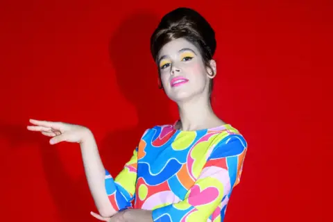 Cosima Ruiz de la Prada, con un peinado voluminoso y maquillaje vibrante, posa con un vestido de estampado psicodélico sobre fondo rojo.