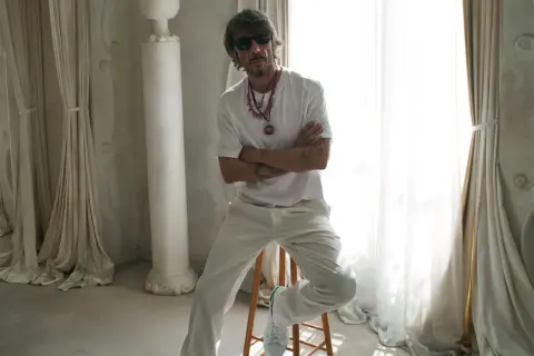 Pierpaolo Piccioli, con gafas de sol y collares llamativos, posa con los brazos cruzados en un entorno elegante con cortinas y columnas.