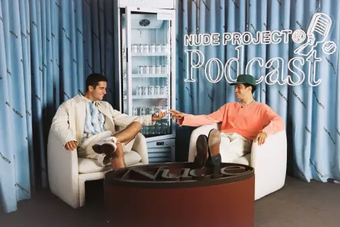 Los fundadores de Nude Project brindan con latas de su bebida, sentados en sillones frente a un cartel de neón del podcast de la marca.