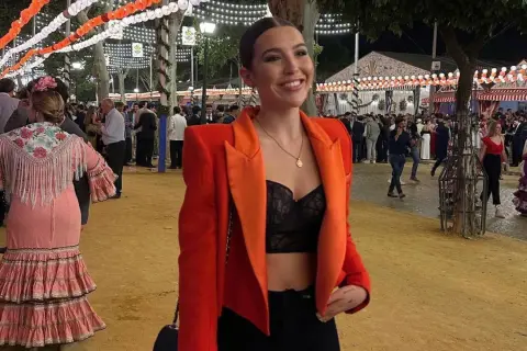 Chica sonriente con chaqueta naranja y top negro, lista para la ropa para ir a la feria de dia. Ambiente festivo con luces y gente.