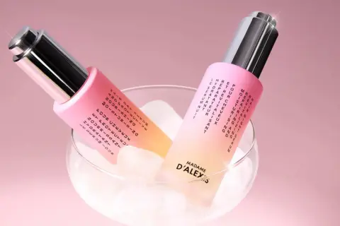 Dos frascos de aceite para pelo, con un degradado rosa y amarillo, reposan sobre cubitos de hielo en una copa.