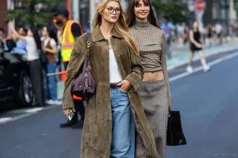 Dos mujeres pasean con un impecable estilo francés. Una lleva un abrigo largo verde oliva, vaqueros y un bolso de Dior. La otra luce un conjunto de cuadros con un top corto y falda.