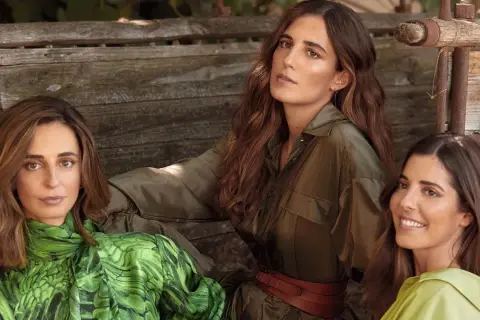 Belen Corsini y dos mujeres más, vestidas con ropa de tonos verdes, posan sentadas en un banco de madera rústico.
