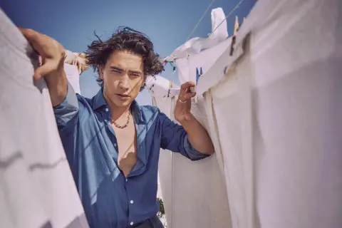 Enzo Vogrincic, con su camisa desabrochada y collar llamativo, posa entre sábanas blancas tendidas al sol.