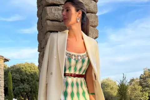 María de la Orden, con un vestido estampado y blazer blanco, posa junto a una pared de piedra bajo un cielo azul.