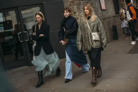 Tres mujeres caminan, mostrando que zapatos se llevan este invierno: botas altas negras, botines marrones y zapatos negros.