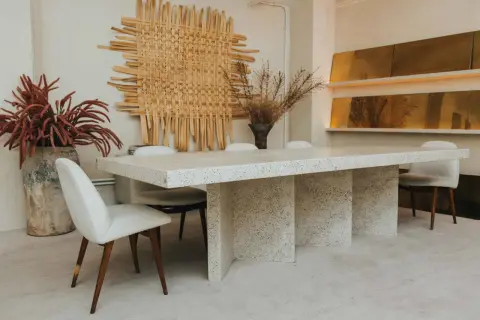 Comedor con mesa de terrazo y sillas tapizadas, arte de pared tejido y jarrones con flores secas. Un ambiente de lujo silencioso.