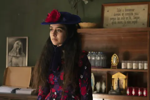 Irene Balmes, con un sombrero azul y un vestido floral, mira pensativa. Detrás, estanterías con objetos religiosos y un cuadro.
