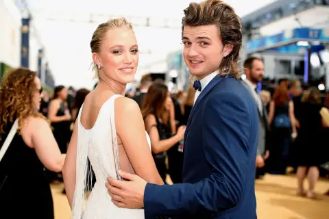 Joe Keery y su pareja, ambos sonriendo a la cámara en una alfombra roja. Ella lleva un vestido blanco con flecos, él un traje azul.