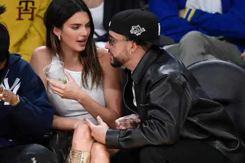 Bad Bunny y Kendall Jenner conversan en un evento deportivo, ella con un vaso en la mano y él con chaqueta de cuero.