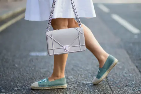 Piernas con un outfit veraniego, falda blanca y un bolso gris, completado con unas cómodas alpargatas de tela azul.