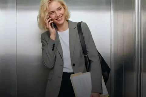 Mujer sonriente en ascensor, con un elegante outfit oficina, hablando por teléfono y cargando carpetas.